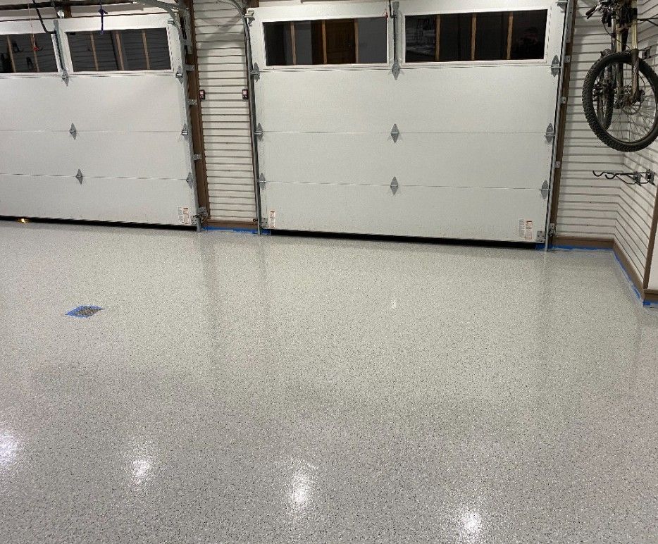 Garage interior with white epoxy flooring speckled with gray. Two closed white garage doors are in the background. A bicycle hangs on the right wall.