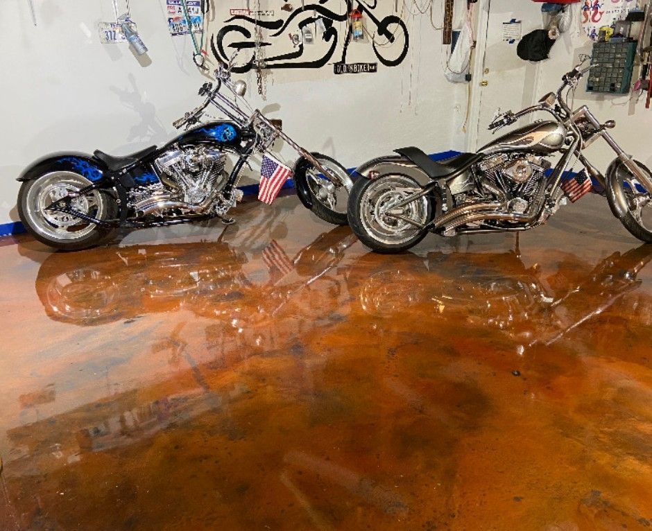 Two custom motorcycles parked on a reflective, metallic-look garage floor. The blue bike is on the left; the silver bike on the right.