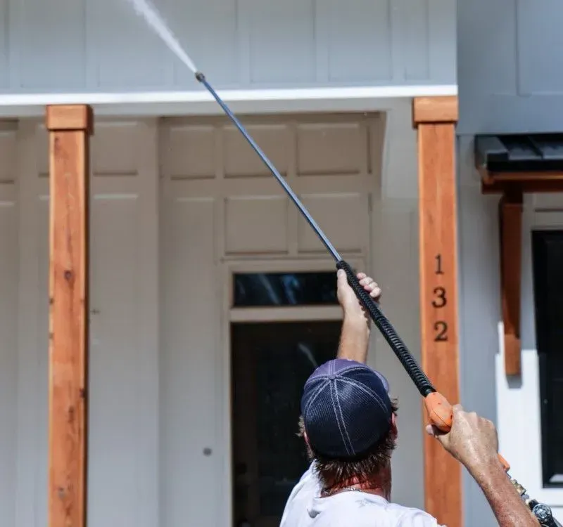 Man pressure washing the exterior of a white house with brown wooden columns.