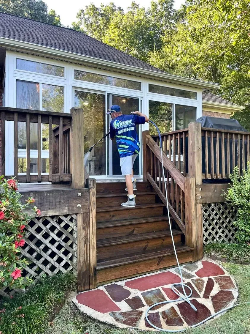 Person power washing a wooden deck with a glass-walled sunroom in the background.