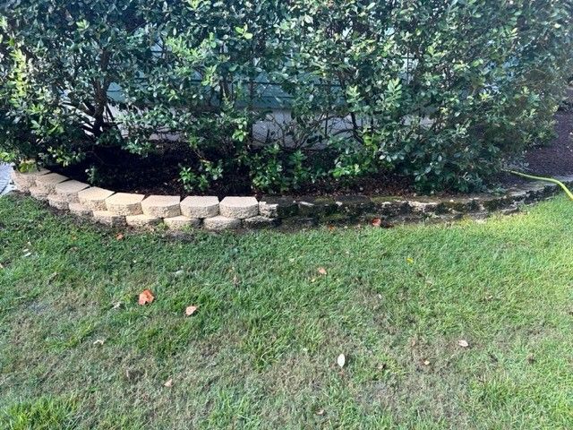 Green grass in front of a curved retaining wall with dark soil and bushes behind it.