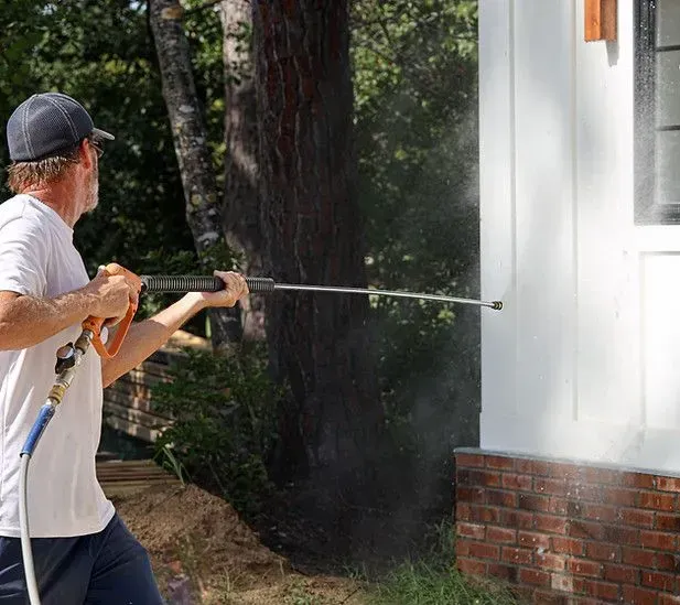 Person pressure washing white siding on a house, water spraying.