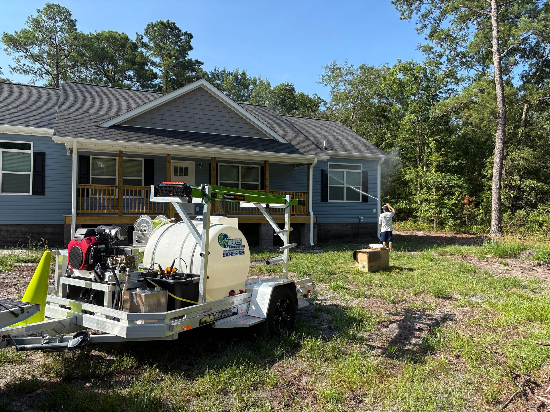 A white trailer with a tank and hose sprays a blue house, person standing nearby, sunny day.