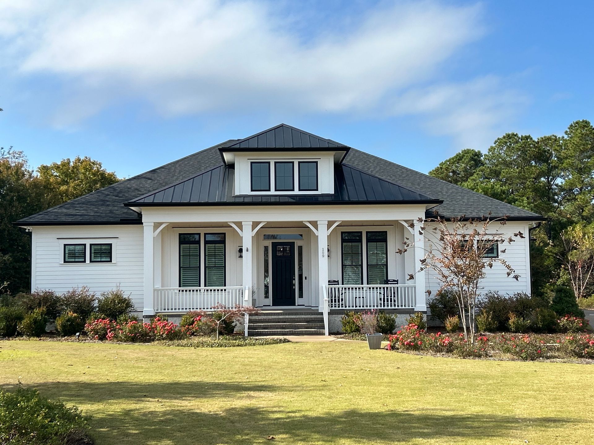 White house with black roof, porch, and rose bushes against a blue sky.