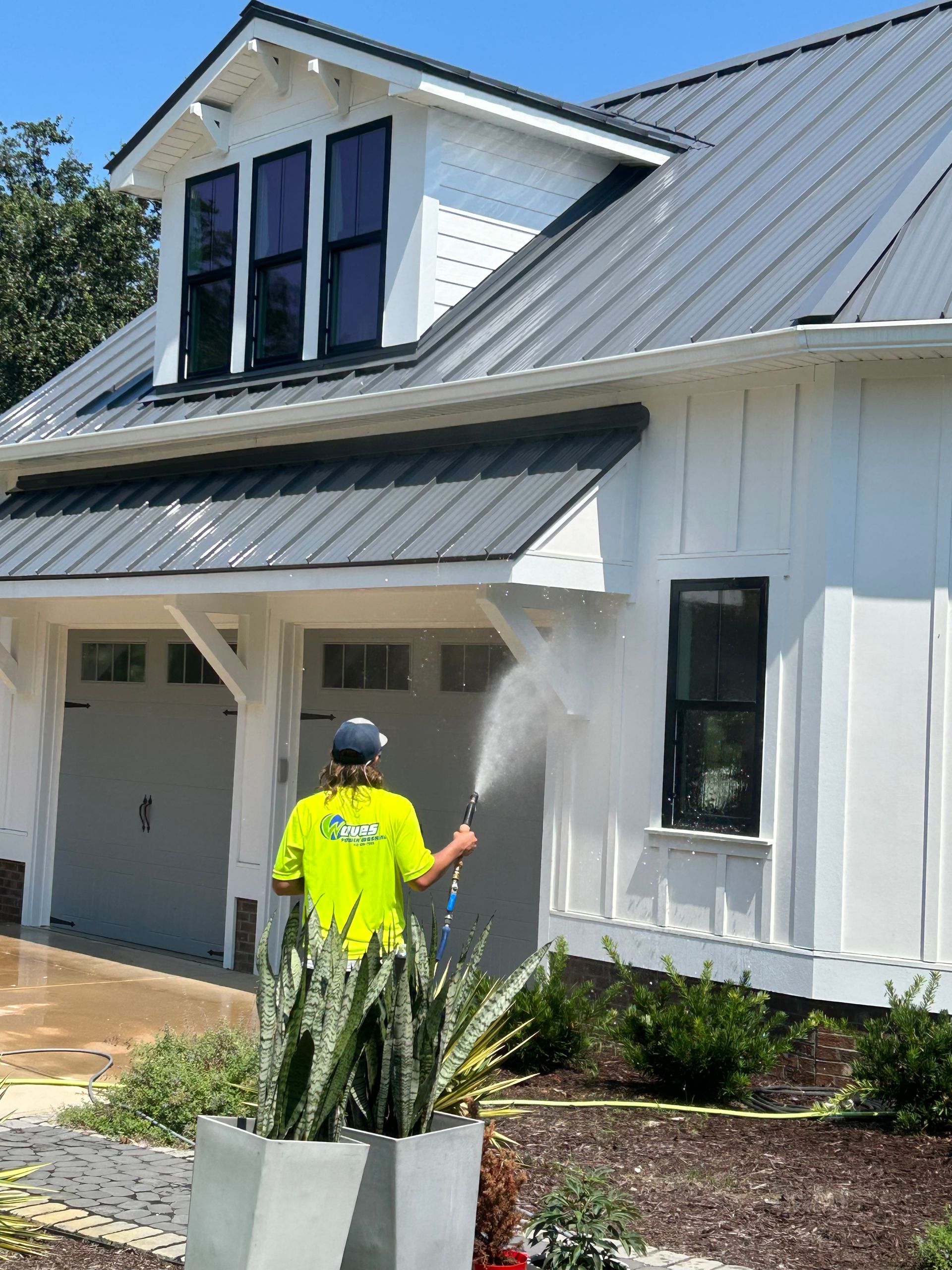 Person in neon shirt pressure washes a white house with a gray roof and black window frames.