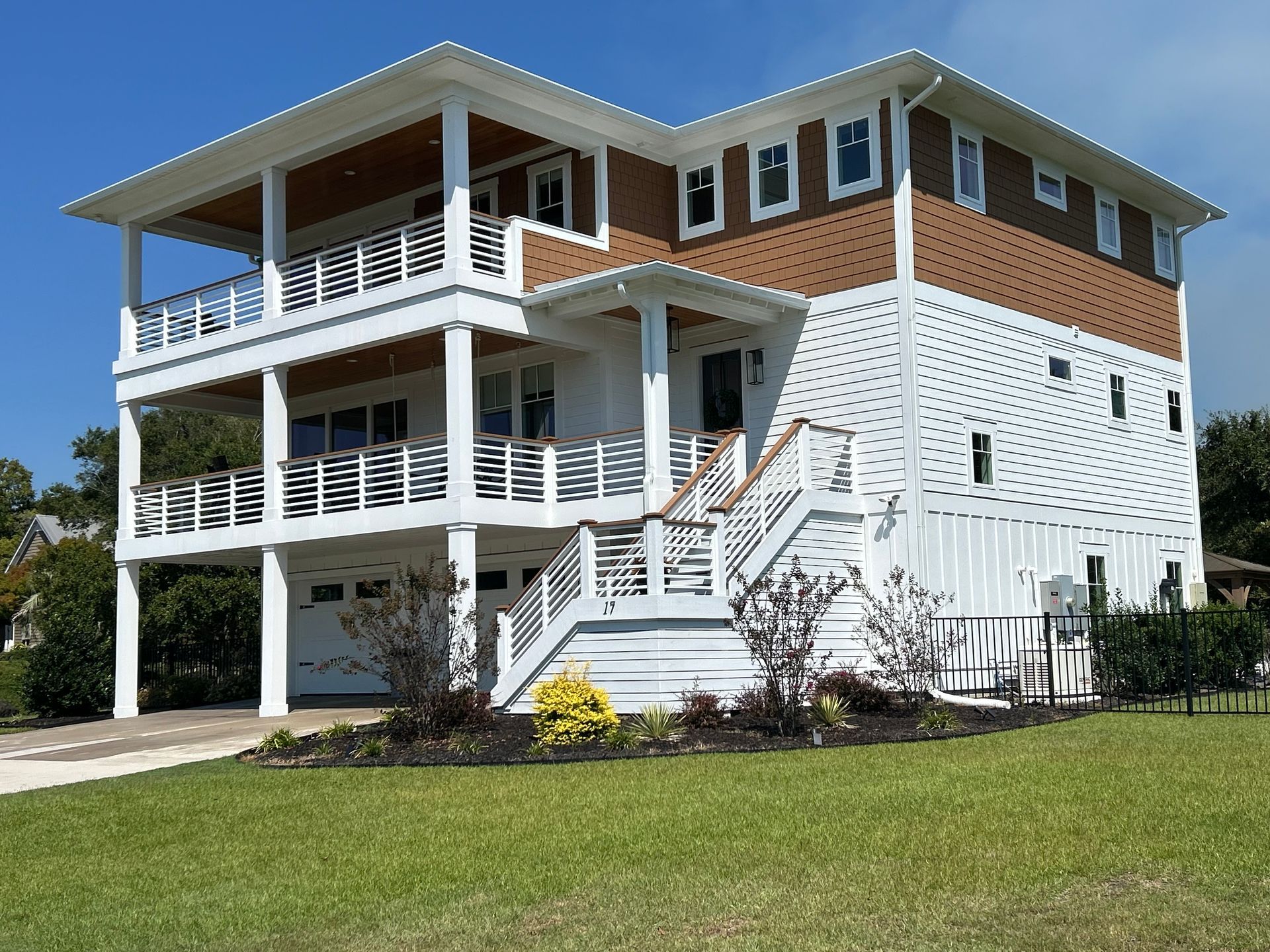 Two-story white and brown beach house with balconies, stairs, and green lawn under a blue sky.