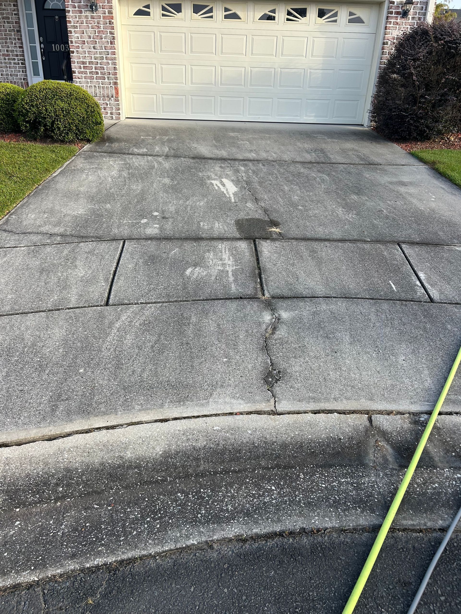 Concrete driveway with cracks leading to a garage. Green grass and shrubs are on either side.