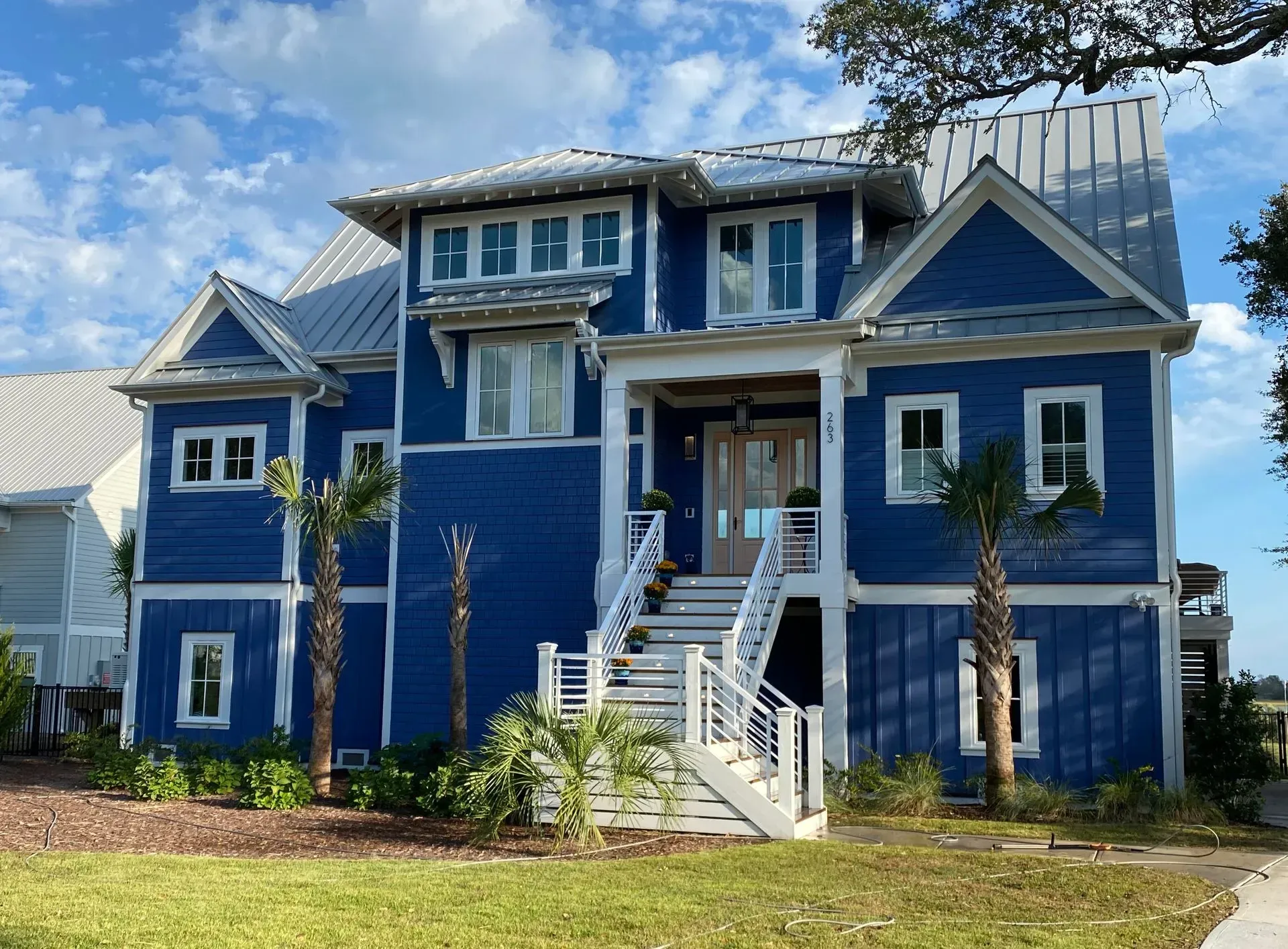 Blue two-story house with white trim, steps, and silver roof. Palm trees in front on a sunny day.