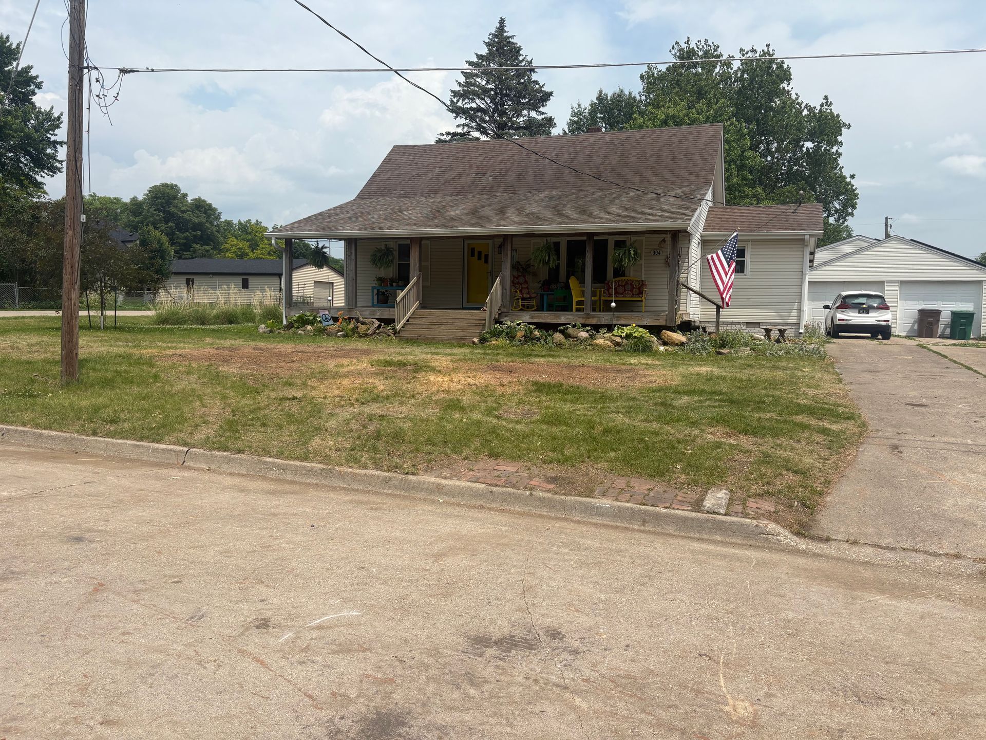 Small, weathered house with porch, American flag, and detached garage on a grassy lot.