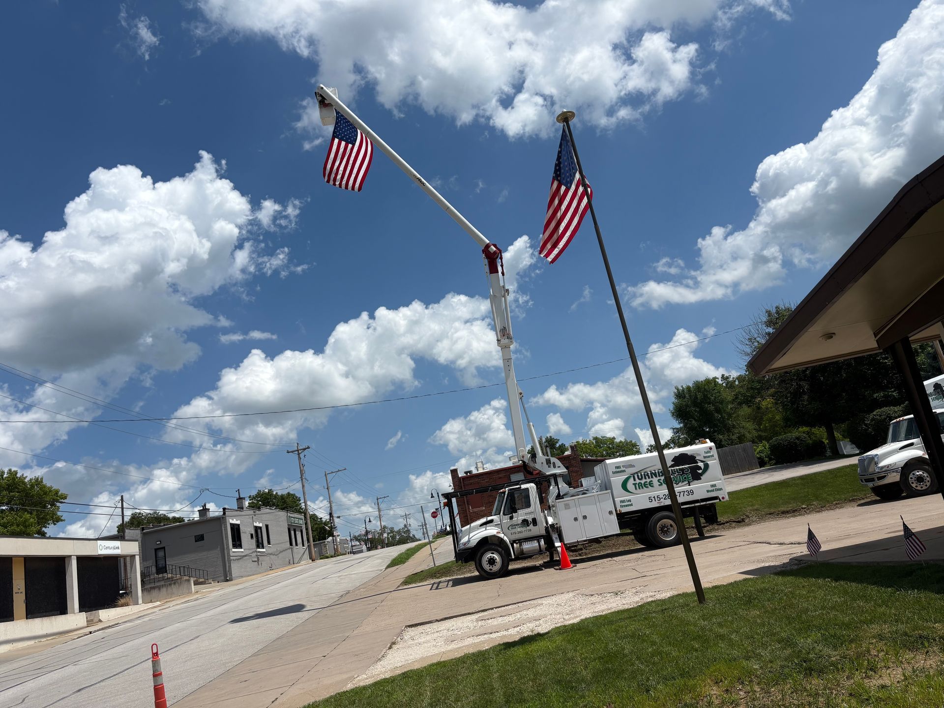 Bucket truck with American flags on a street, under a blue sky with clouds, roadside view.