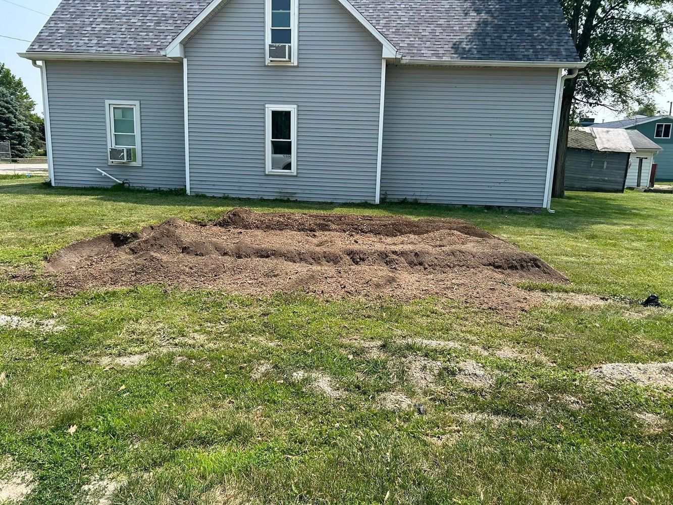 Pile of dirt in a grassy yard in front of a gray house.