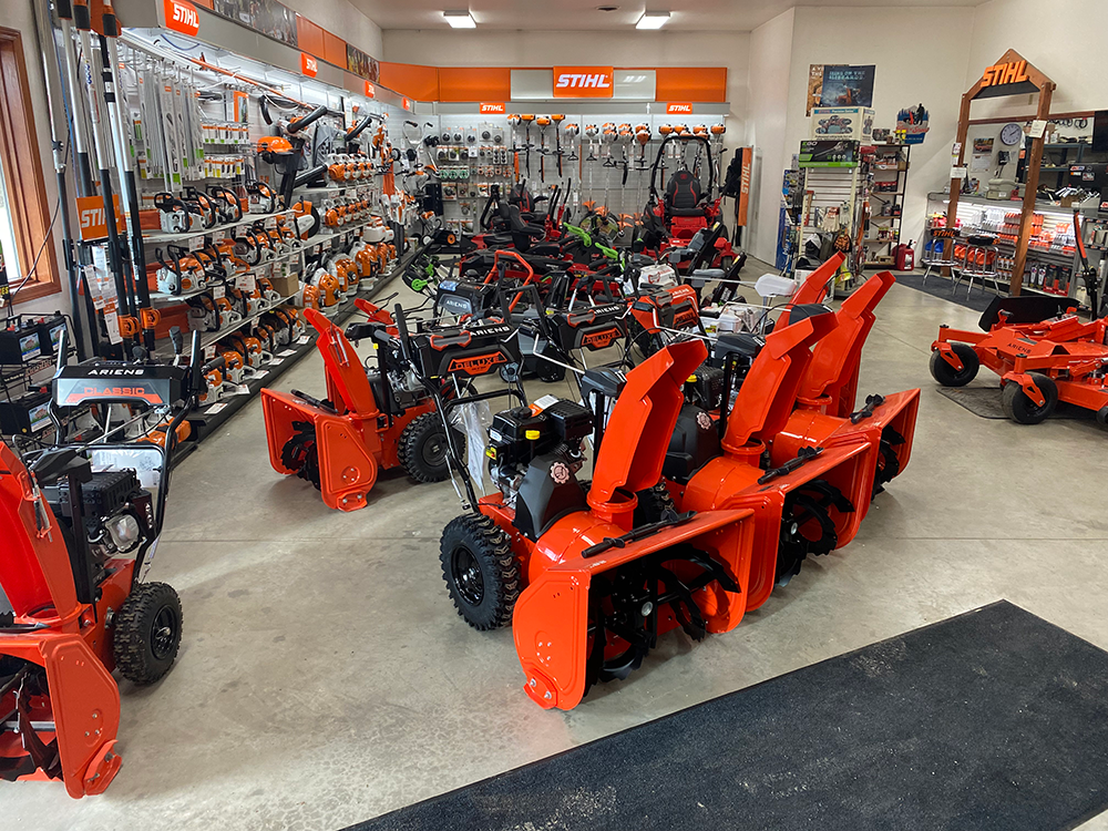 A showroom interior displays multiple orange snow blowers arranged on a concrete floor, with STIHL products on walls.