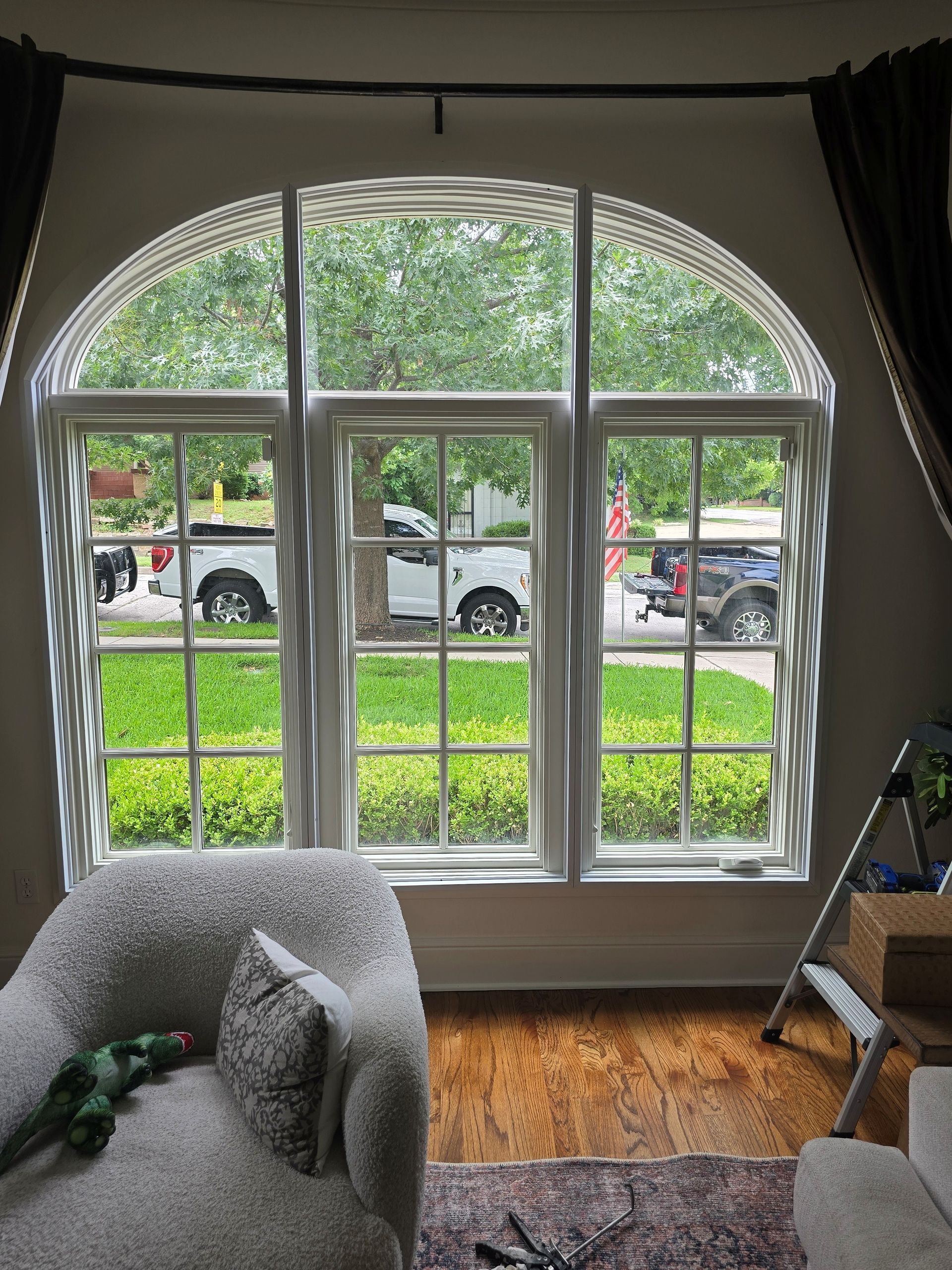 Arched window with three panes, looking out at a street with vehicles and greenery.  An armchair, rug, and wooden floor are visible.