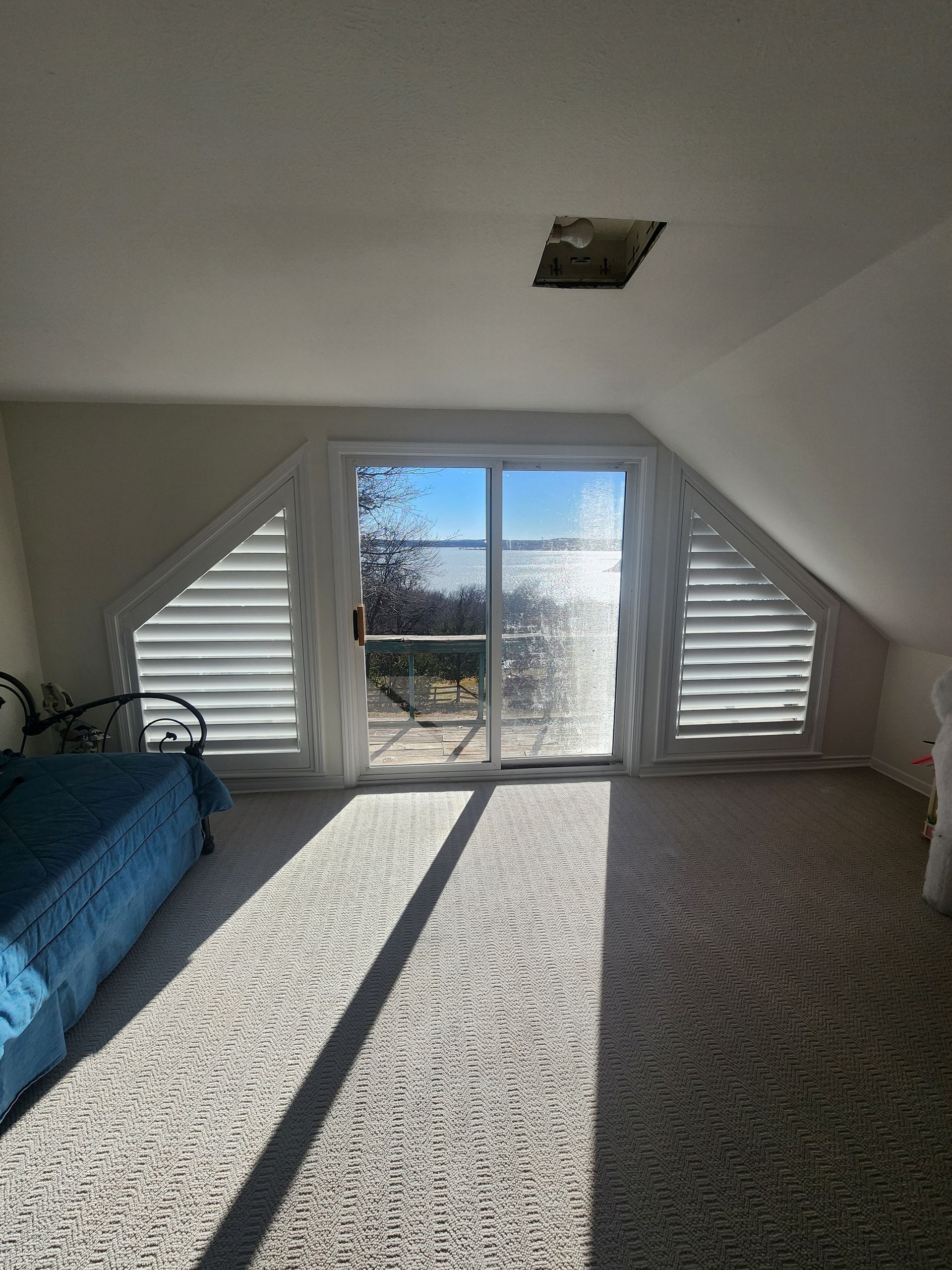 Sloped-ceiling bedroom with large window and slatted shutters. Sunlight streams across the carpet.