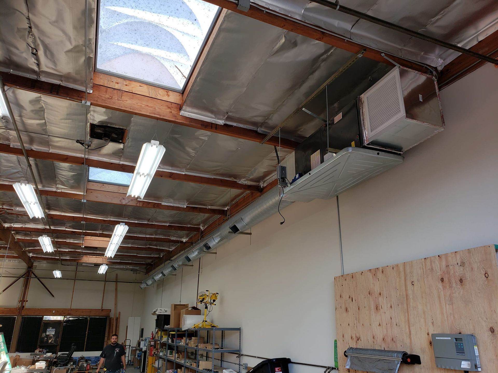 Interior view of a workshop with exposed rafters, ductwork, and fluorescent lighting. A person is visible in the foreground.