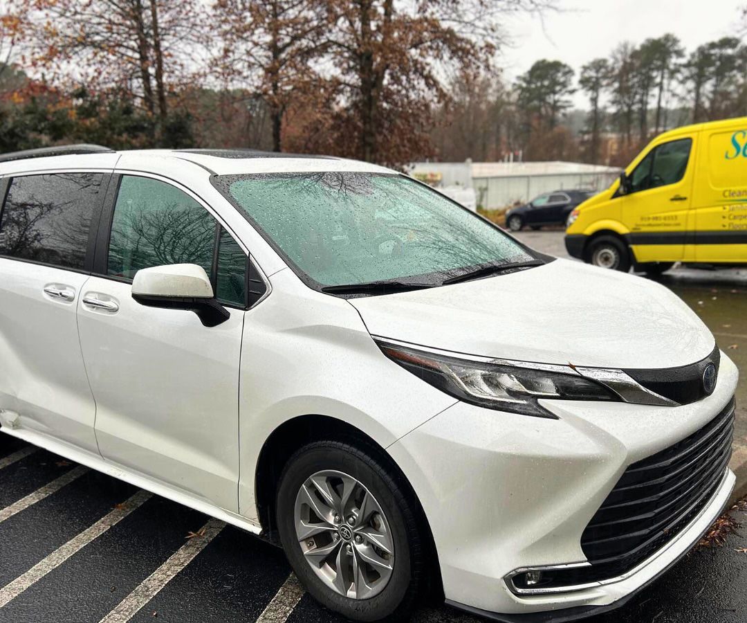 White Toyota Sienna minivan parked outside on a wet day, yellow van in the background.
