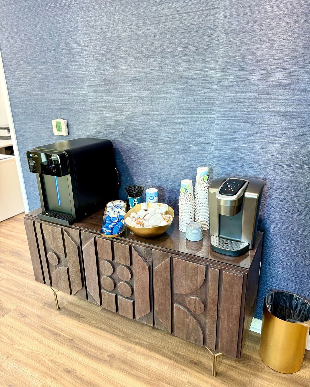 Coffee station with machine, cups, snacks, on a wood cabinet against a blue textured wall, and a gold trash can.