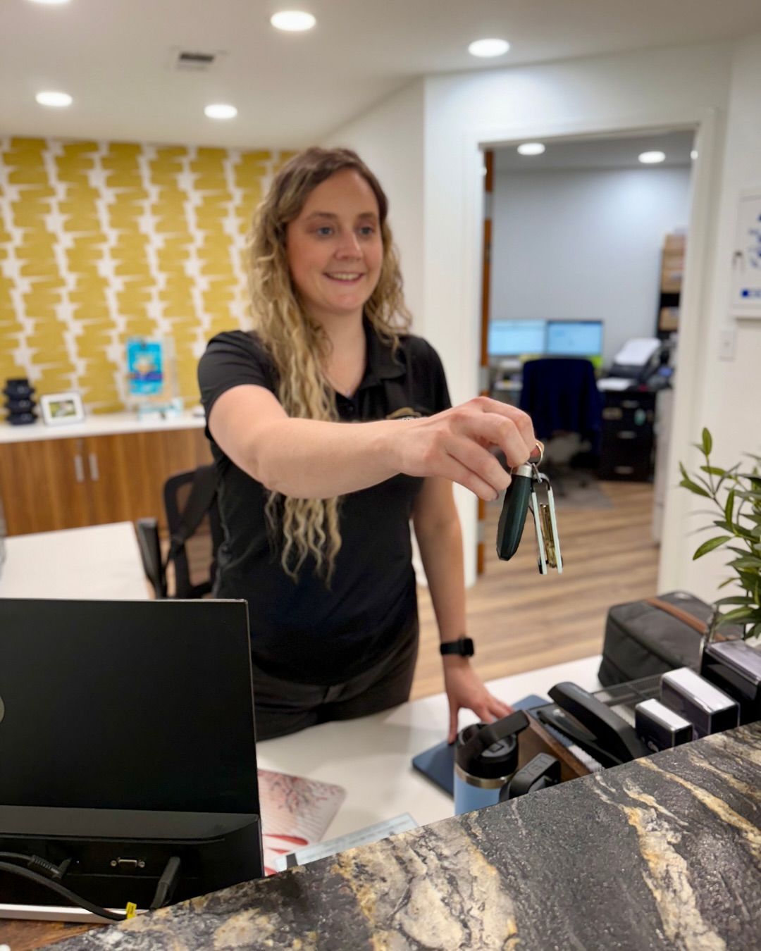 Woman in black shirt hands car keys at a front desk in a light-filled office setting.