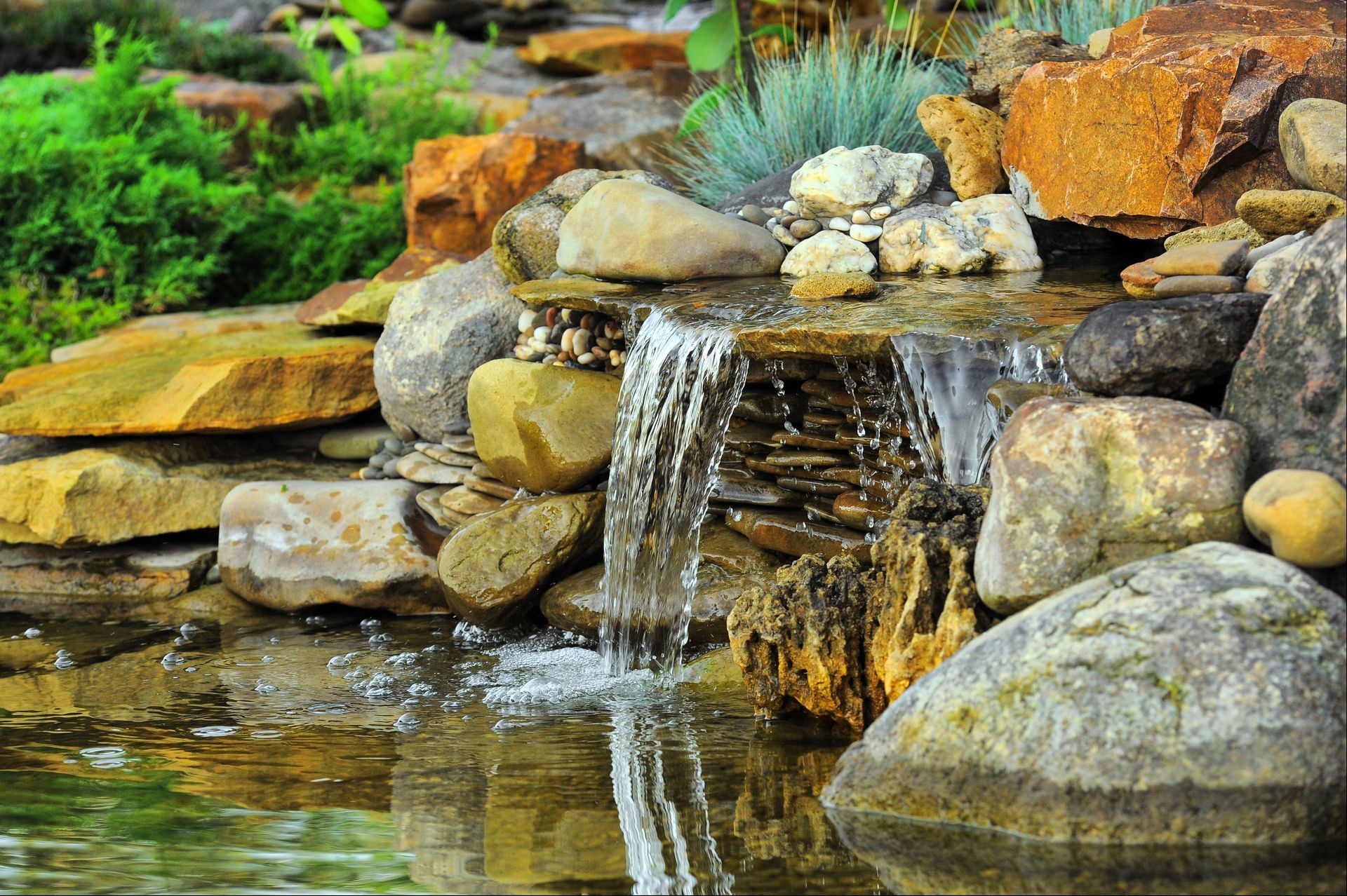 Small waterfall cascading over rocks into a pond. Brown and gray stones surround water and greenery.