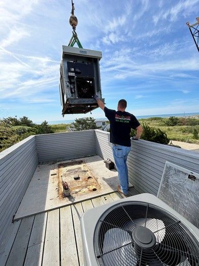 A worker watches as a crane lifts an HVAC unit off a roof. Beach and blue sky in background.