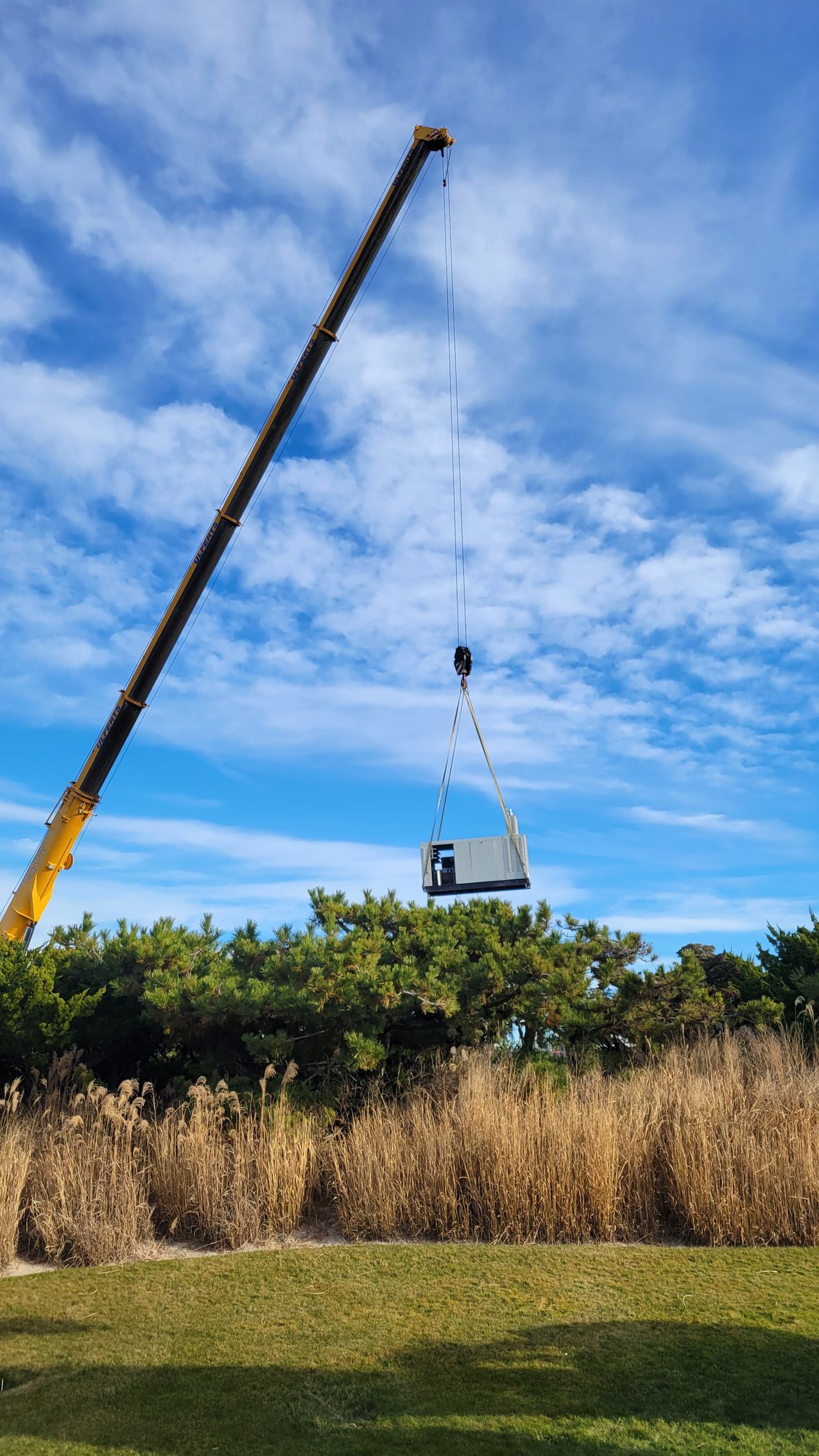 A crane lifts a gray rectangular structure above green bushes under a blue sky.
