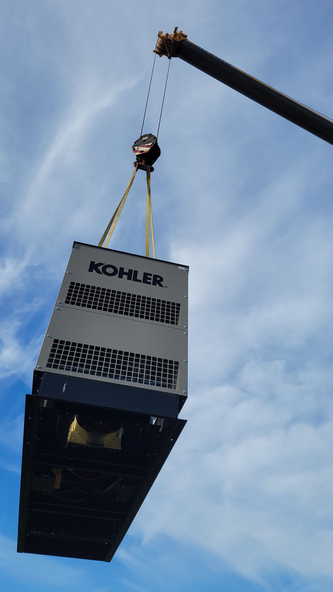 A crane lifts a Kohler generator outdoors against a cloudy sky.