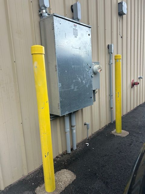 Yellow bollards protect an electrical box on a building's exterior. Gray conduits and beige siding are also visible.