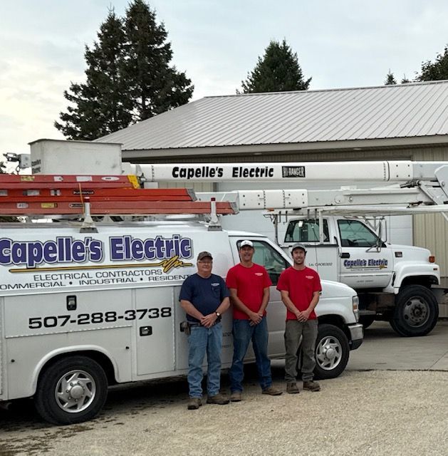 Three electricians in front of Capelle's Electric trucks. Two wear red shirts, one blue. Building behind.
