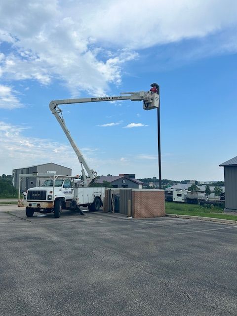 A worker in a bucket truck repairs a tall black pole under a cloudy blue sky.