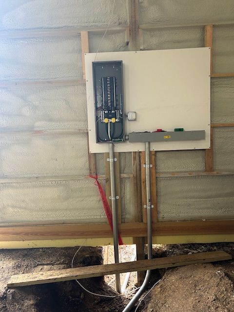 Electrical panel mounted on a building wall, with metal conduit and wires exposed near a dirt trench.