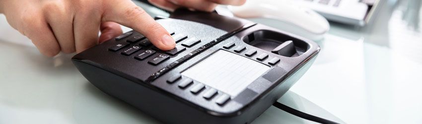 Hands dialing a black office desk phone beside a laptop on a white desk
