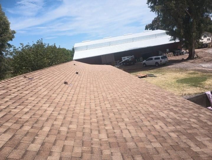Rooftop with tan shingles leading to a white metal building and parked vehicles under trees