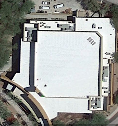 Aerial view of a large white-roofed building with surrounding trees and parked vehicles.