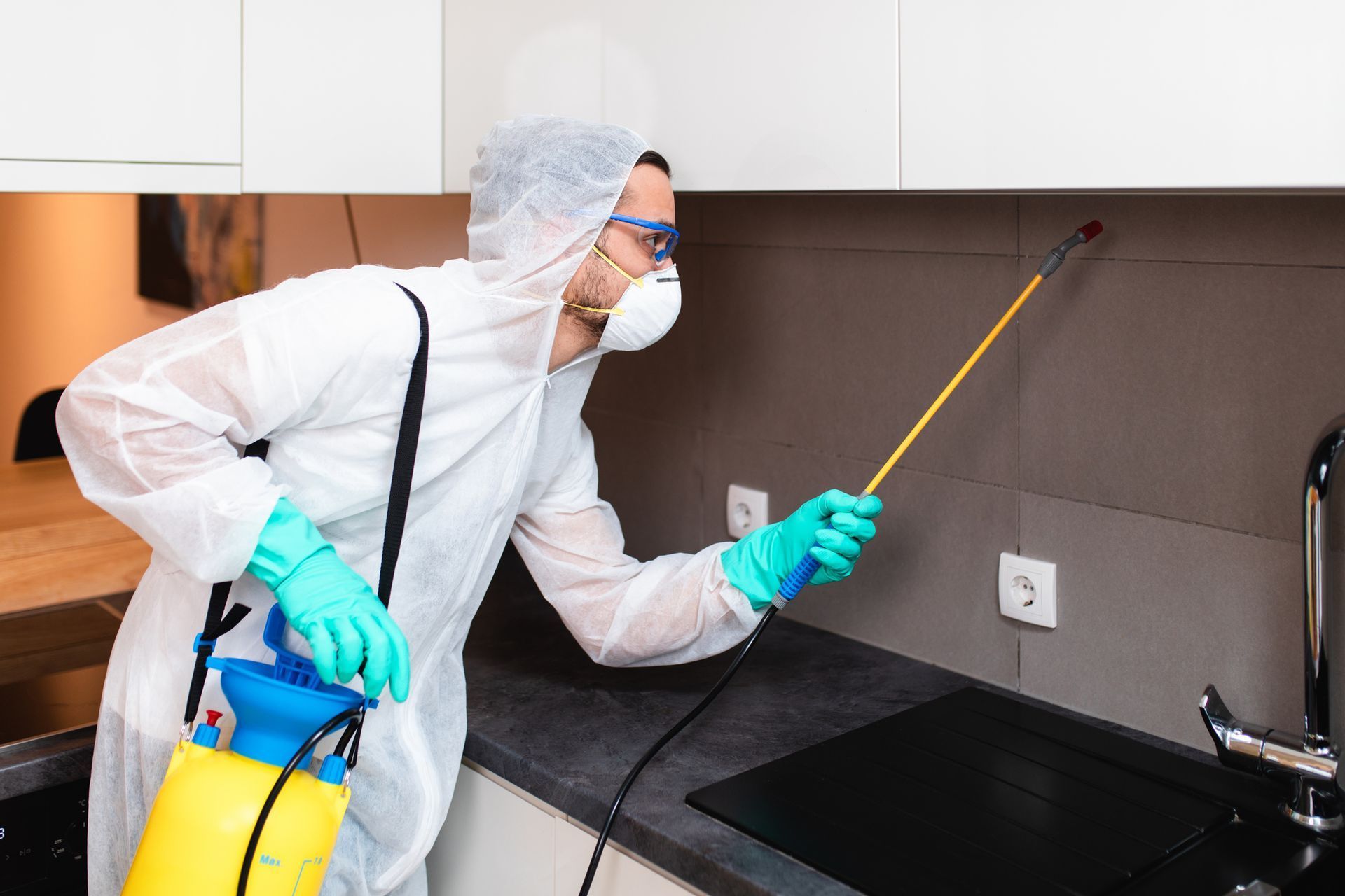 A professional in a white protective suit, mask, and gloves uses a yellow handheld sprayer to treat a kitchen wall.