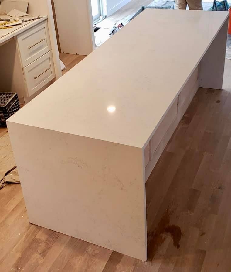 Large, white countertop island with light wood cabinets, set on a hardwood floor, inside a room under construction.