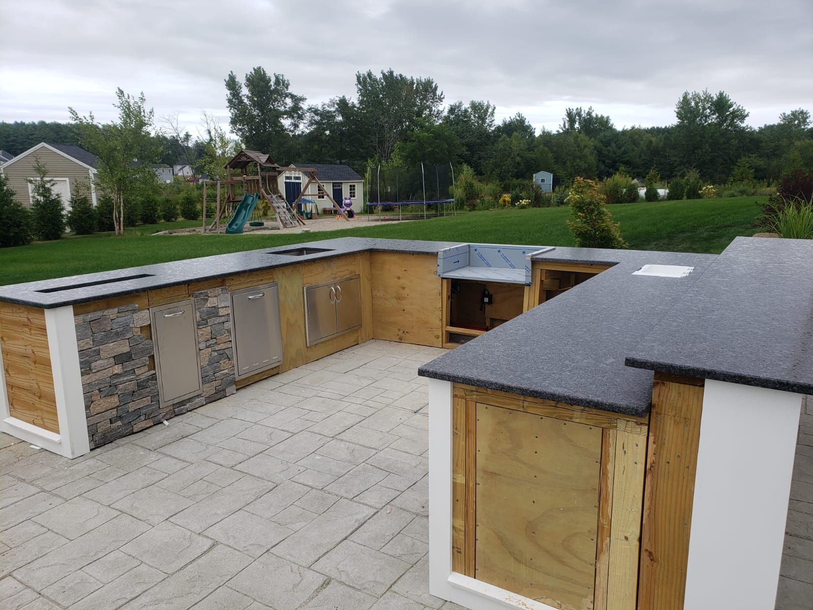 Outdoor kitchen under construction, with granite countertops, wooden cabinets, stone and stainless steel accents, on a patio.