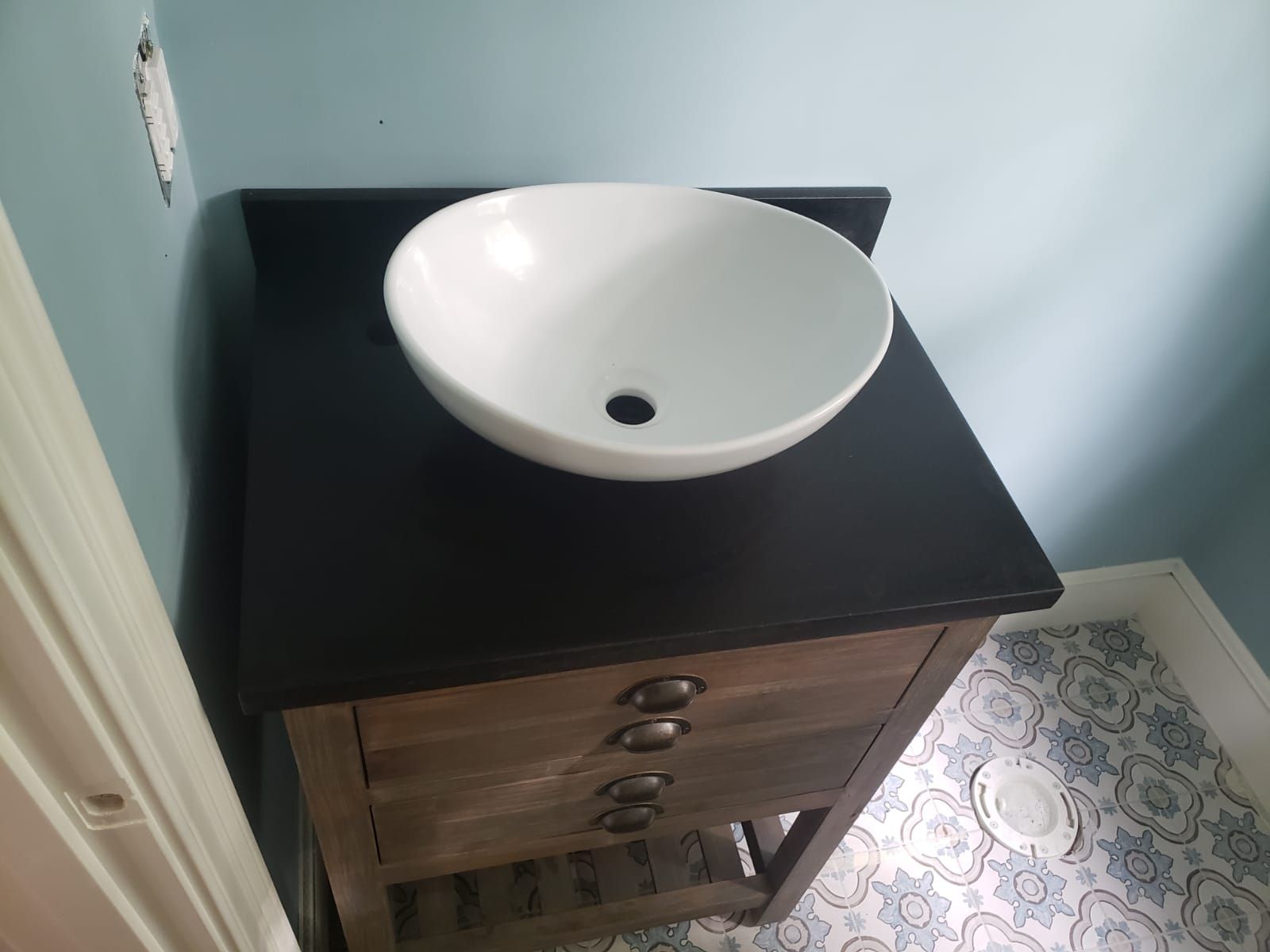 Wooden bathroom vanity with a black countertop and white vessel sink. Blue walls and patterned floor.