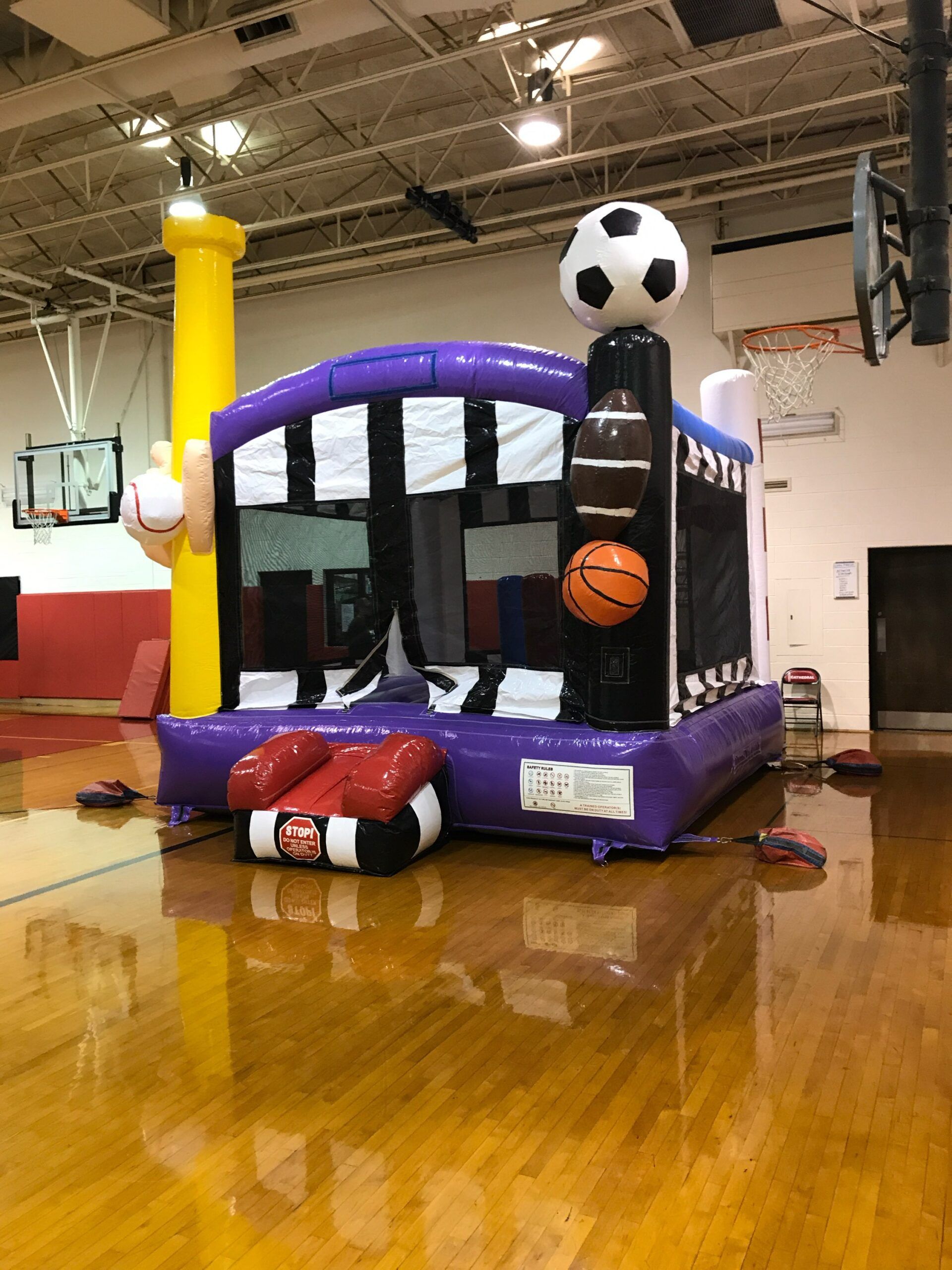A purple bouncy house with a soccer ball , basketball , and football on top of it in a gym.