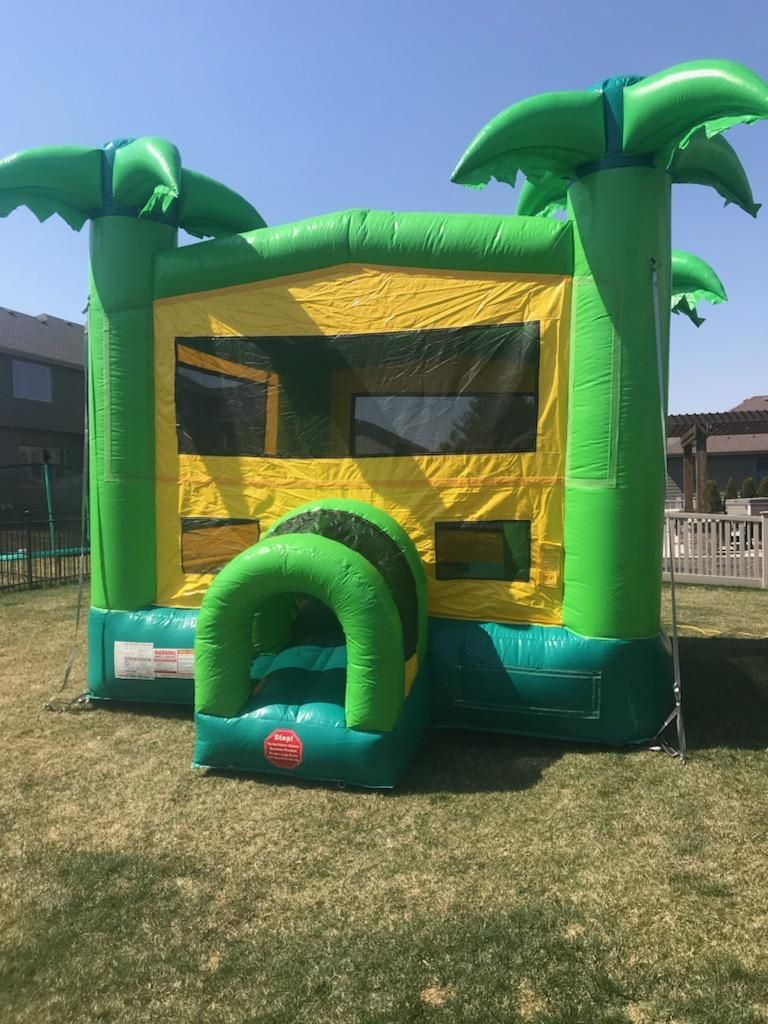 A green and yellow bouncy house with palm trees on top is sitting on top of a lush green field.
