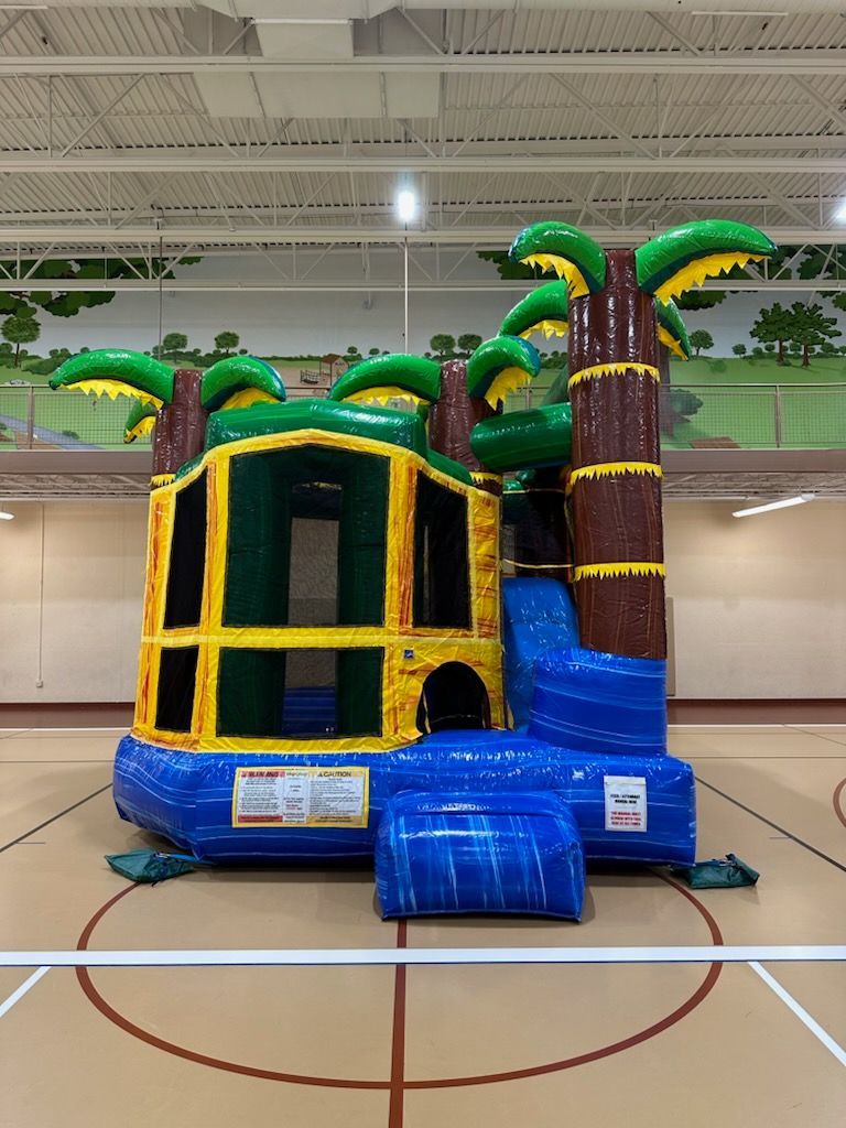A bouncy house with palm trees and a slide in a gym.
