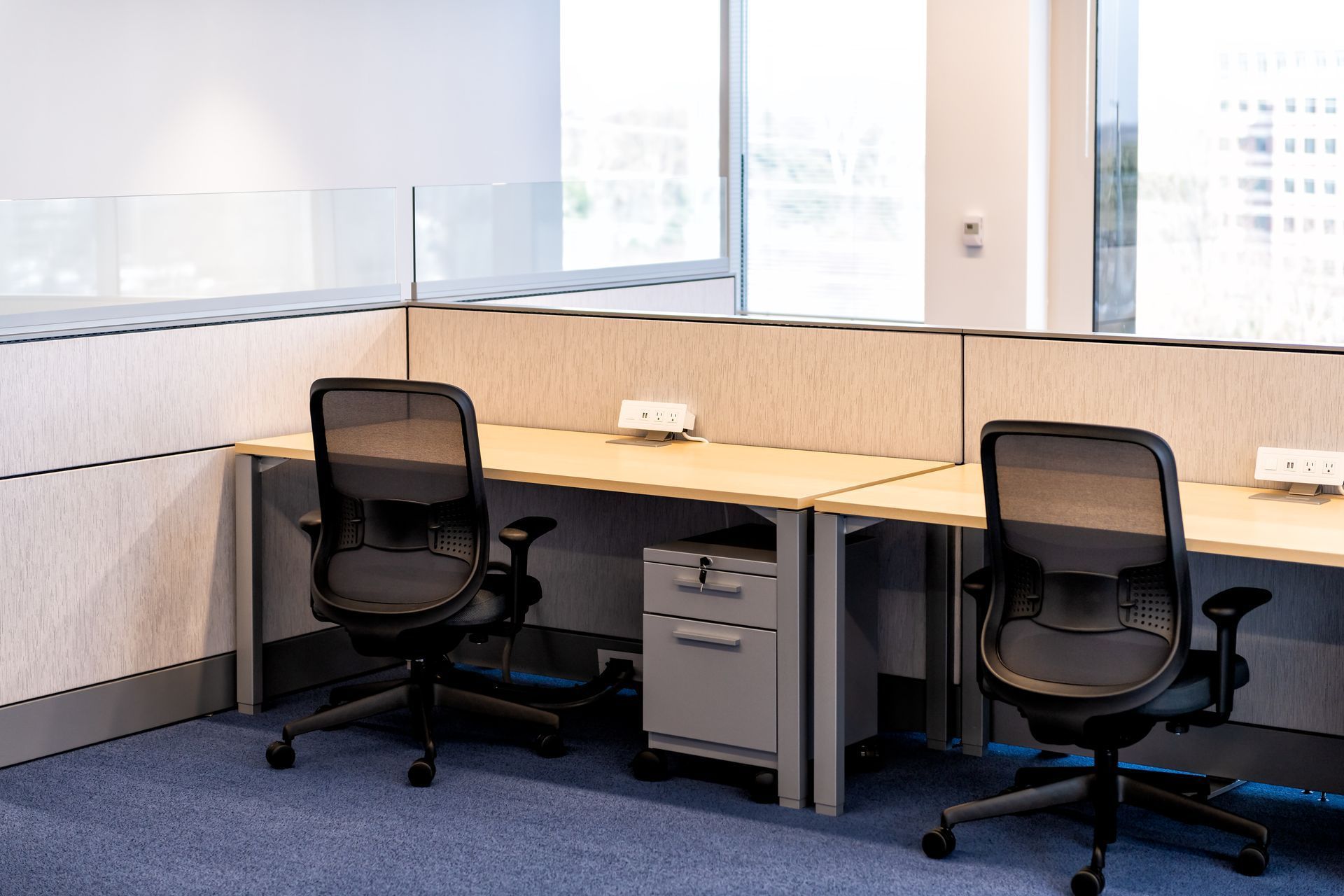 Empty office cubicle with two black chairs, wooden desks, and large windows.