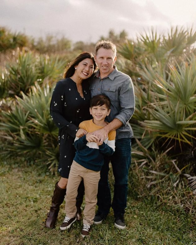 Family of three smiling outdoors among tall green plants, with parents hugging a child.
