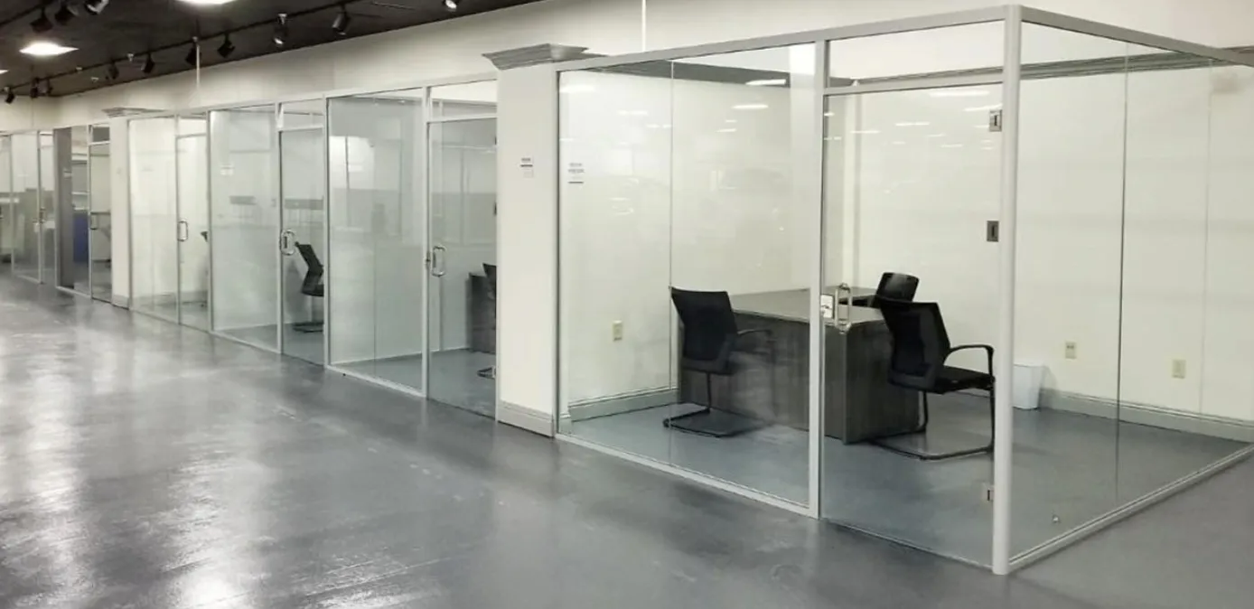 Glass-walled office pods in a bright open workspace, with desks and chairs inside.