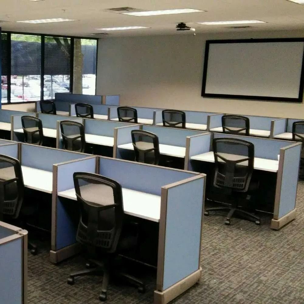 Empty office training room with rows of blue cubicles, swivel chairs, and a projector screen.