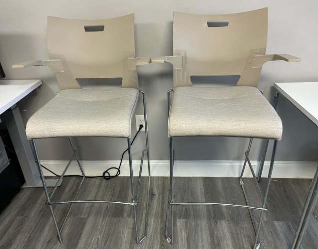 Two beige bar stools with metal legs beside white desks on a gray wood floor.