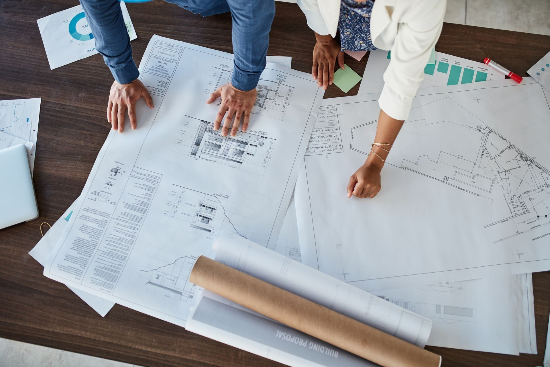 People reviewing architectural blueprints on a desk with rolled plans and a laptop