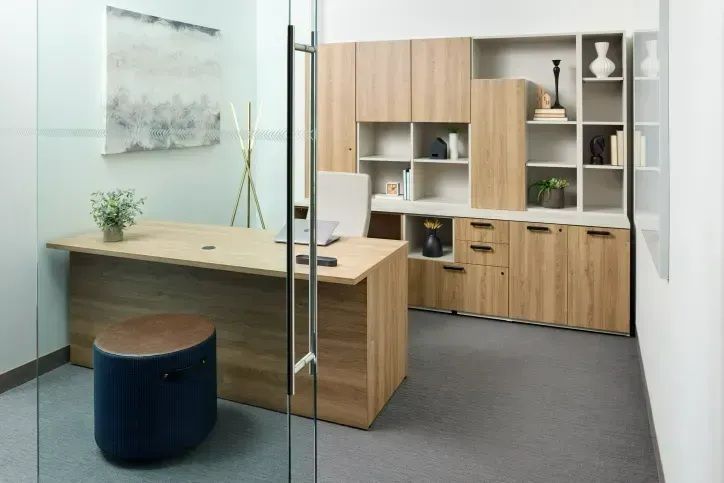 Modern office with glass wall, wooden desk, storage cabinets, and a blue stool in the foreground