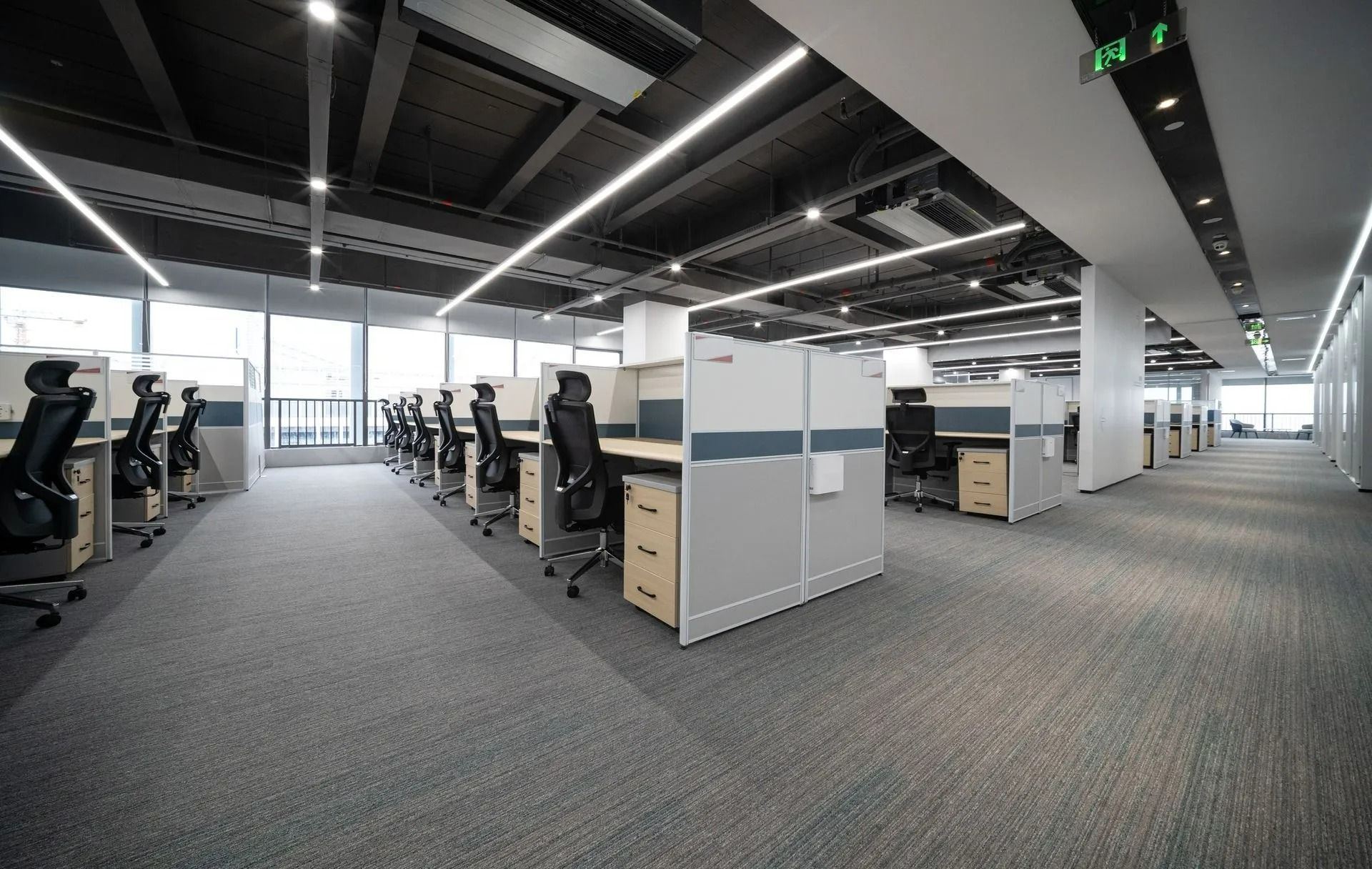 Modern open-plan office with rows of desks, monitors, and carpeted flooring under bright ceiling lights