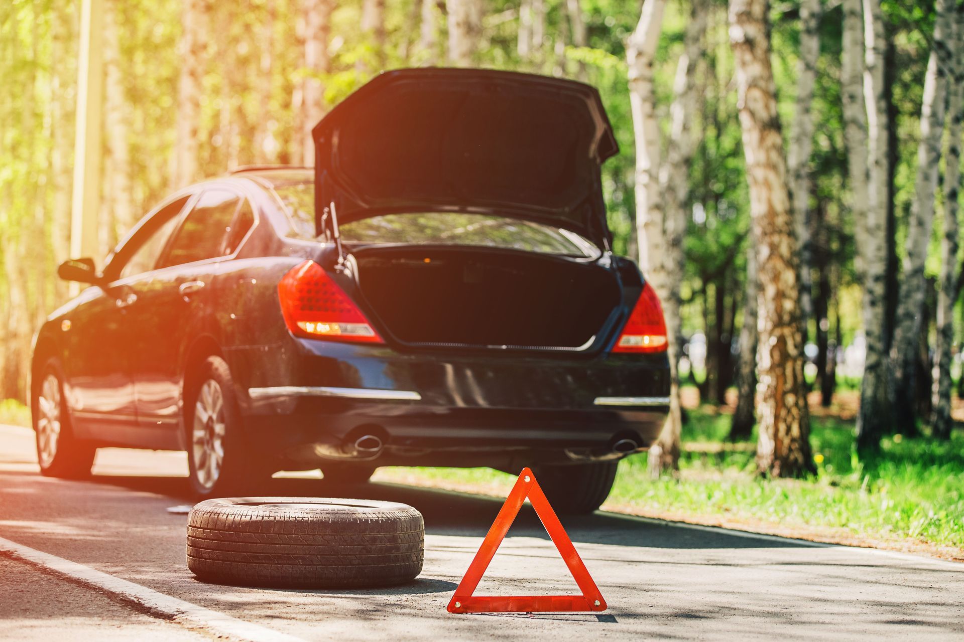 Car with a flat tire and open trunk on the side of a road, with an orange triangle warning sign.