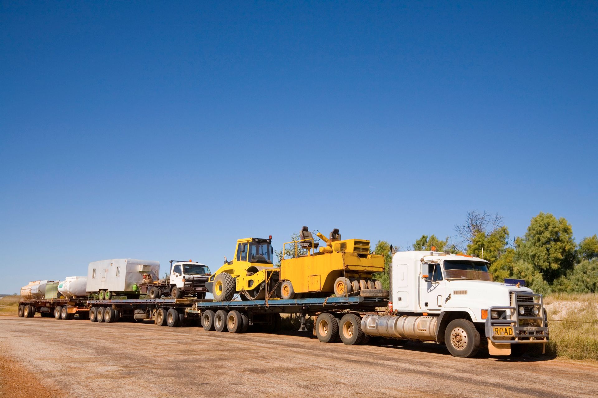A long truck hauling construction equipment on a dirt road under a clear blue sky.